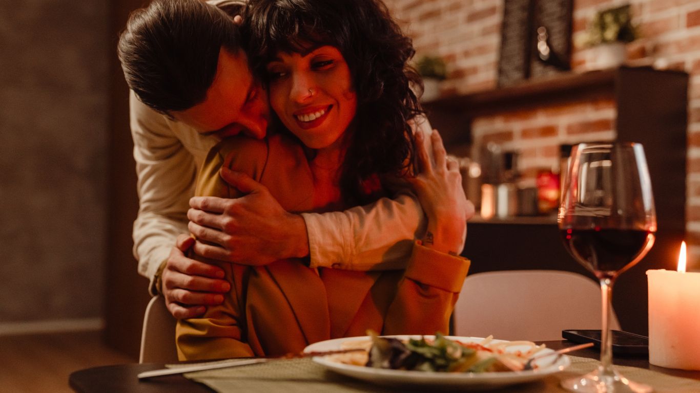 couple cuddling next to a table with a candle lit meal, plate of food and glass of red wine celebrating valentines Cambridge