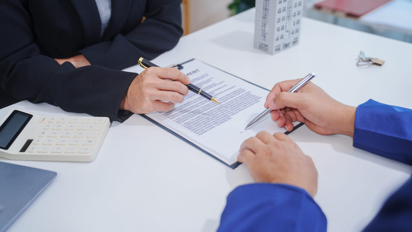 A person signing an agreement with another person pointing to the document on the other side of a table