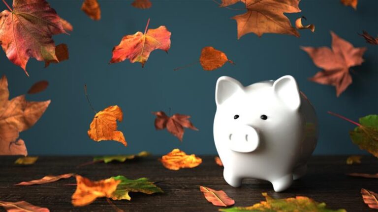 A white piggy bank on a wooden table with falling orange leaves around it